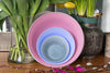 Set of nesting handblown glass bowls with a Pale Rose large, Cornflower Blue medium, and Light Grey small propped up against vessels with spring flowers in them. Made in the USA from Serve Kindness.