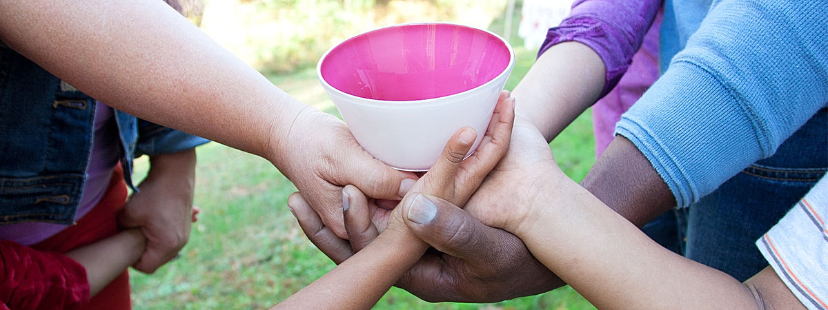 A multiracial families' hands stack on top of each other holding a pink kindness bowl, symbolizing that each glass bowl purchase donates to kindness charities, Mosesian Center of the Arts and 50 Legs.